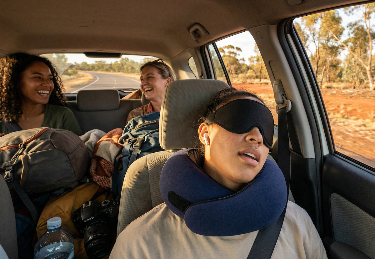 Traveler resting comfortably with neck pillow and eye mask on Australian road trip, friends enjoying the journey in background